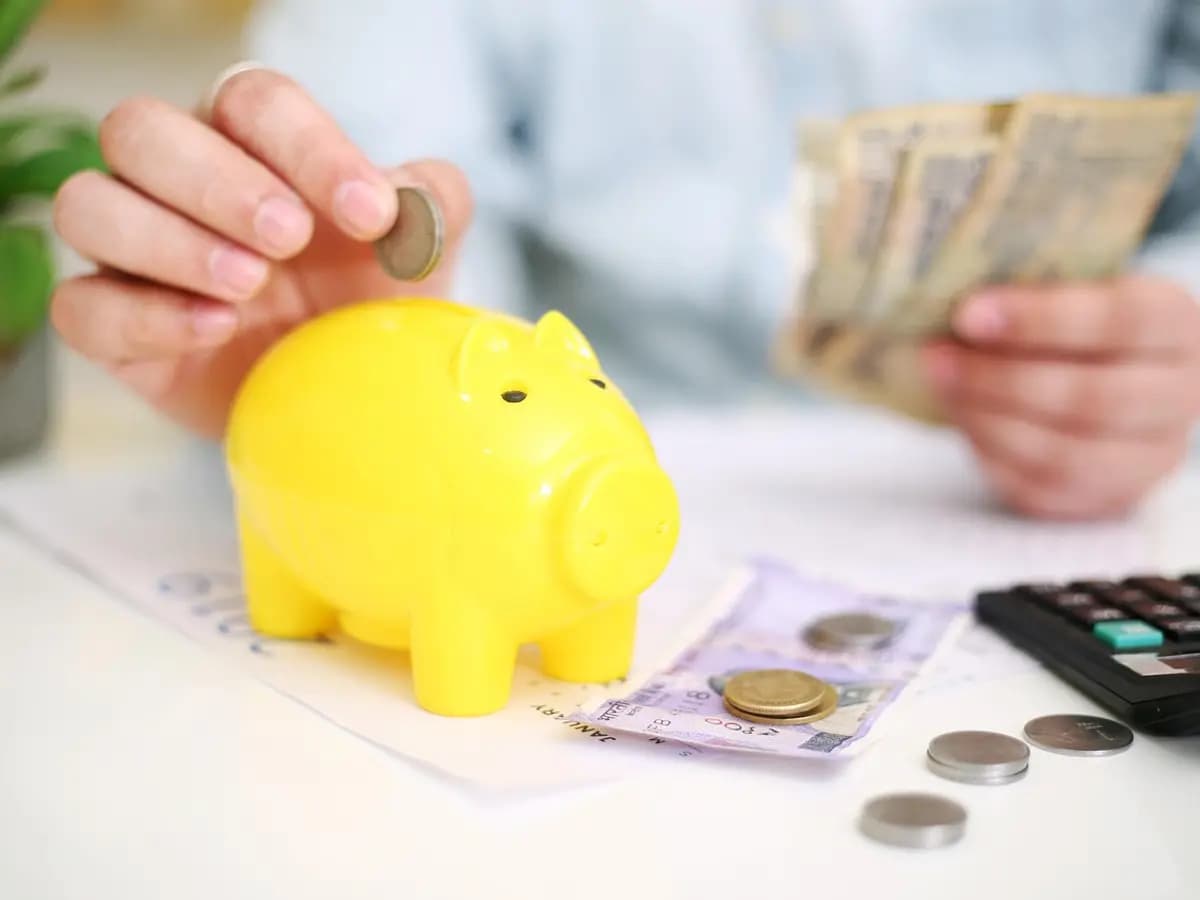 A hand dropping a coin into a yellow piggy bank beside rupee notes, coins and a calculator