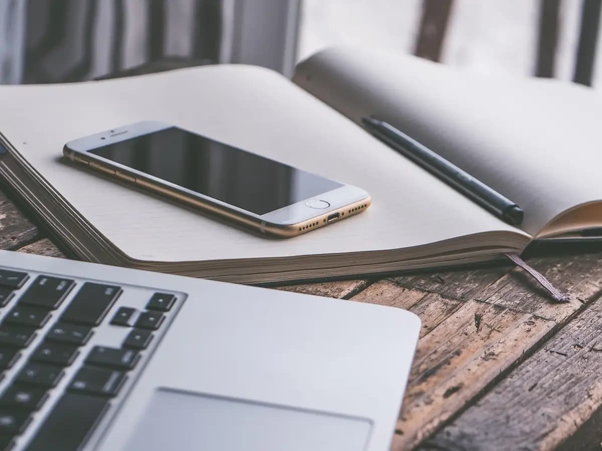 A smartphone lying on an open notebook next to a laptop keyboard on a wooden desk